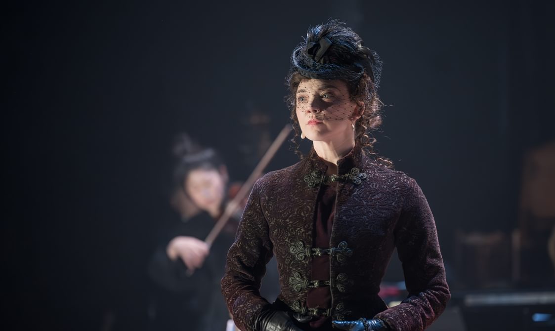 A woman in a detailed black coat with black leather gloves and a black fascinator is stood with her hands clasped in front of her. In her hands she is holding a small red velvet pouch. She has a serious look on her face. Behind her, a woman in all black is playing the violin.