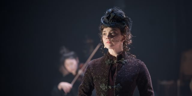 A woman in a detailed black coat with black leather gloves and a black fascinator is stood with her hands clasped in front of her. In her hands she is holding a small red velvet pouch. She has a serious look on her face. Behind her, a woman in all black is playing the violin.