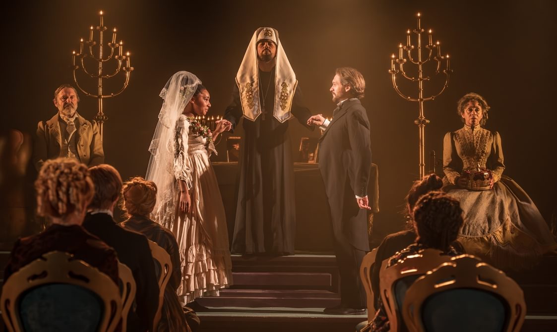 A wedding scene. A Russian Orthodox minister is stood between the bride and groom, who is wearing a black tailed suit. Her wedding dress is extravagant, with ruffles and flowers and a beautiful tiered lace veil. There are golden candelabras either side of them with a man sat on the left and a woman on the right, both with slightly solemn expressions. There are several members of the congregation facing the ceremony.