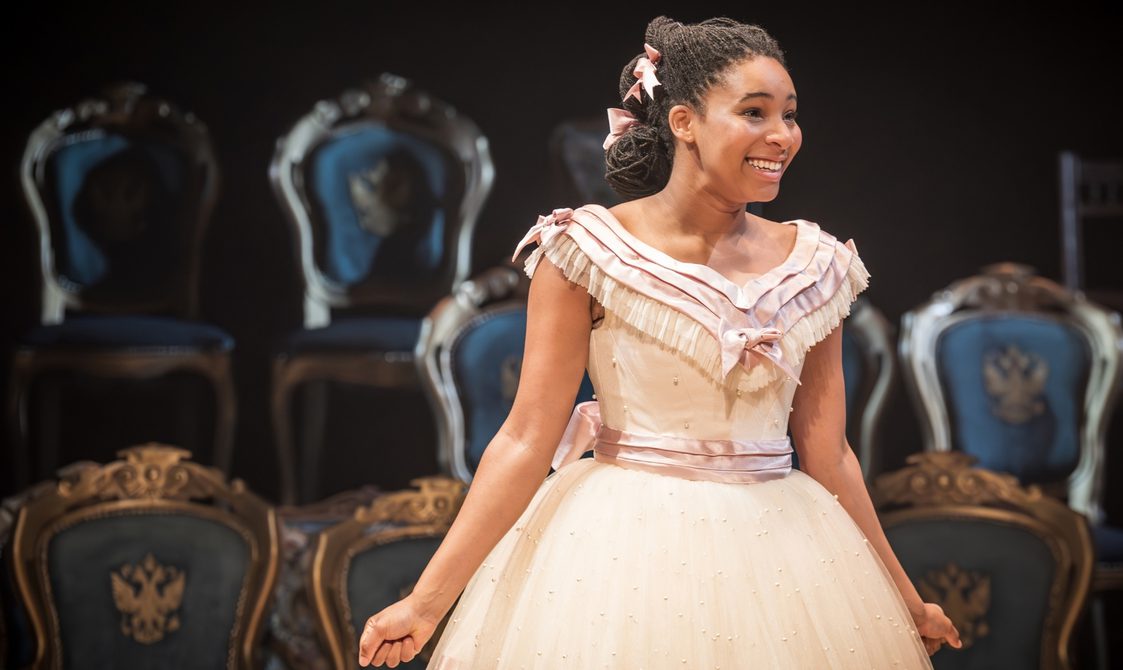 A young woman is stood excitedly with a bright smile on her face. She has pink silk bows in her hair and is wearing a pretty dress in the same colour, which is adorned with pearls and ribbons.