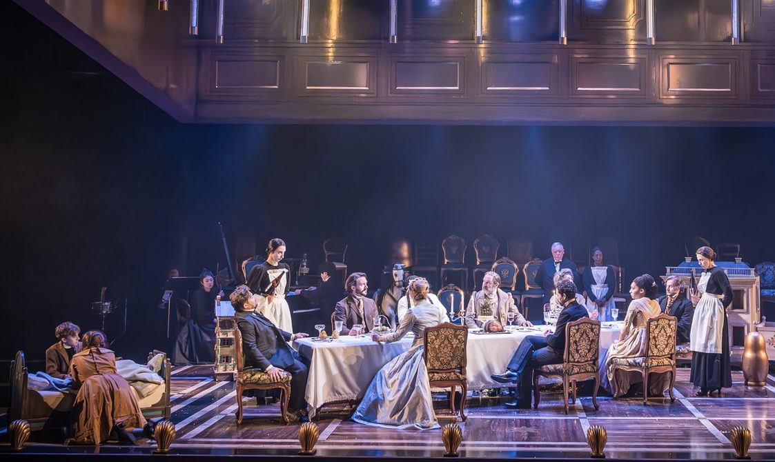 A dinner scene with many performers with serious expressions. The long dining table is dressed with a white tablecloth and the performers are sat on ornate vintage dining chairs. On the left, two children are leaning on a small bed.