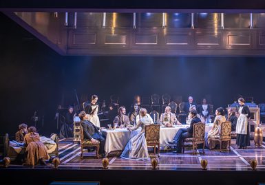 A dinner scene with many performers with serious expressions. The long dining table is dressed with a white tablecloth and the performers are sat on ornate vintage dining chairs. On the left, two children are leaning on a small bed.
