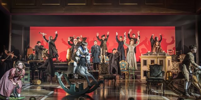A large group of performers on stage. Stood slightly raised at the back, many characters have their arms raised above their heads and their mouths agape. They look panicked. At the front of the stage, three children are each on rocking horses, all with determined looks on their faces.