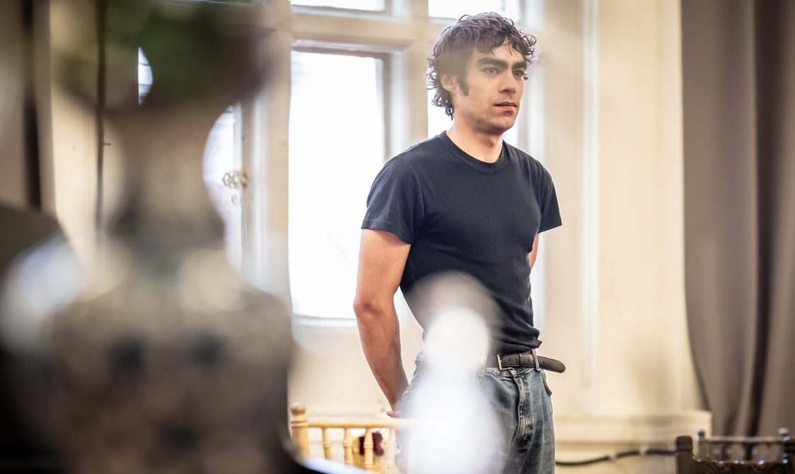 A man with dark hair is stood with his hand behind his back and a neutral expression on his face. He is wearing a black t-shirt and blue jeans.