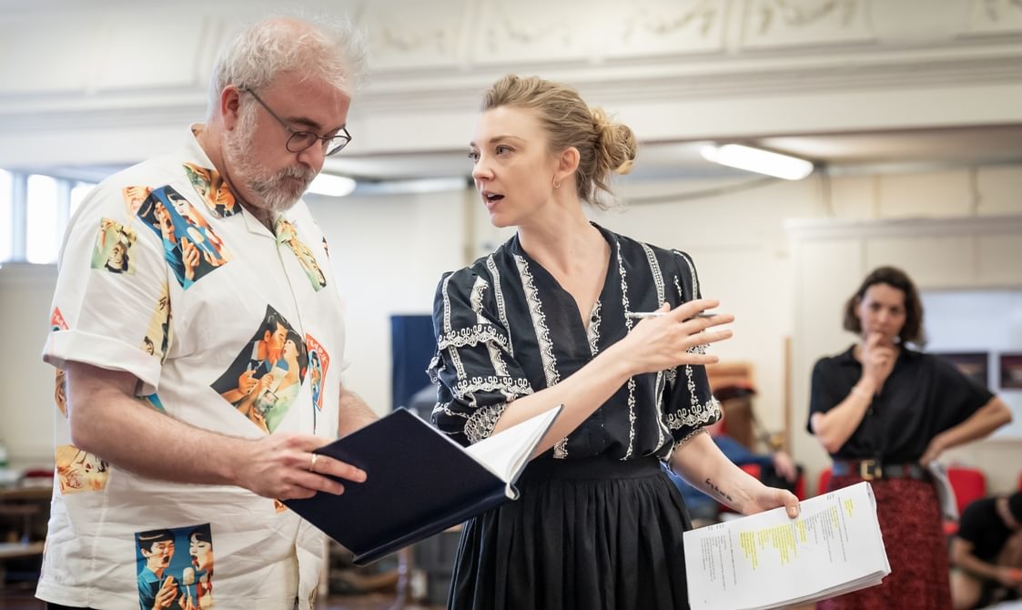 A man with white hair and a beard is reading a notebook which he is holding. He is wearing glasses and a white shirt with various colourful portraits. Next to him, there is a woman with blonde hair talking to him. She is wearing a black blouse with white detailing and a black skirt. She is holding a script with a lot of text highlighted in one hand and a pen in the other. In the background, there is a woman wearing a black top and long red skirt looking at them with one hand on her hip and the other resting on her chin, as if in thought.