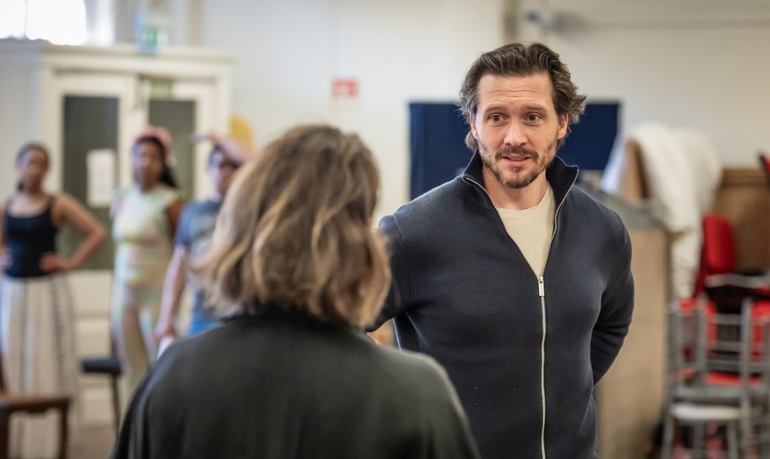 A man with brown hair and a beard is stood facing a woman who has her back to the camera. He is wearing a dark grey zip-up jumper with a white shirt underneath. He has a neutral expression.