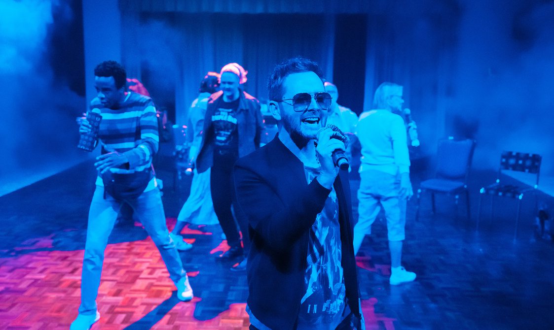 A man wearing aviator sunglasses and a casual blazer over a graphic t-shirt is singing happily into a microphone. Behind him, other performers are also singing into microphones. The lighting is indigo and there is faint haze all around the space.