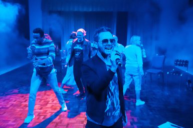 A man wearing aviator sunglasses and a casual blazer over a graphic t-shirt is singing happily into a microphone. Behind him, other performers are also singing into microphones. The lighting is indigo and there is faint haze all around the space.