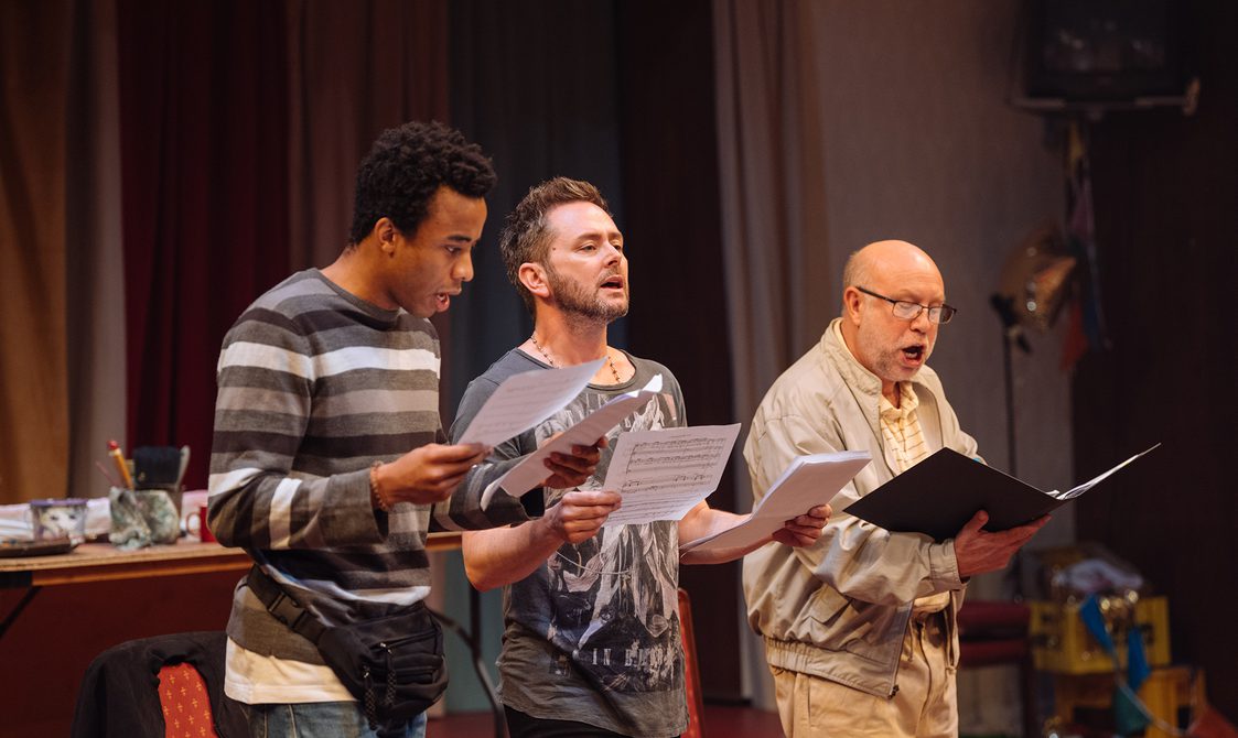 Three performers are stood reading from sheet music and singing. On the left, a young man is wearing a grey striped jumper with a black bum bag around his waist. In the middle, a bearded man is wearing a grey graphic t-shirt. On the right, another man is wearing reading glasses and a beige bomber jacket over a pale yellow check shirt.