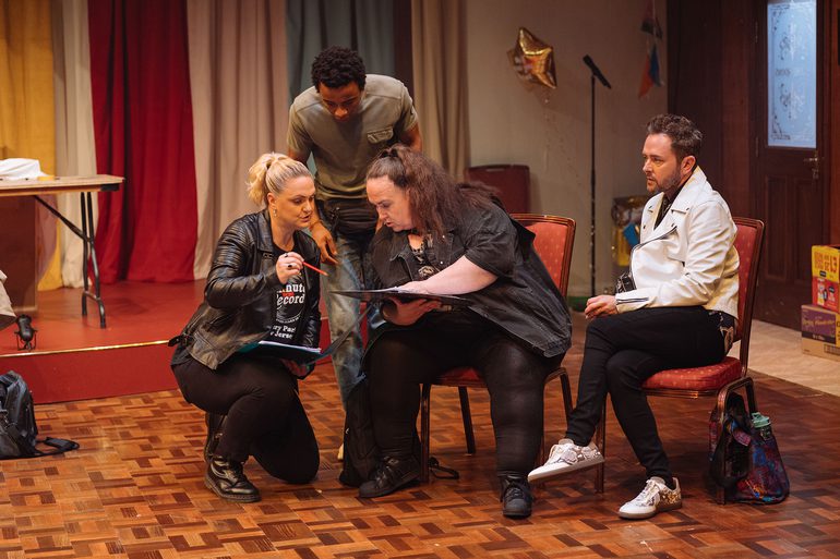 Four performers are gathered in a rehearsal room with colourful curtains and a gold foil star balloon. On the left, a woman with blonde hair, another woman with brunette hair and a young man are all looking at a folder and conversing. A bearded man on the right is staring at them intensely.
