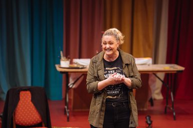 A woman with her blonde hair in a ponytail with an orange pencil tucked through it is smiling excitedly. Her hands are clasped in front of her. She is wearing a casual khaki jacket over a black graphic t-shirt. In the background, there is a table with art supplies in front of swathes of colourful curtains.