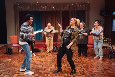A group of performers are stood in a semi-circle, singing gloriously while reading from folders. In the centre, a woman with blonde hair is using a pencil like a conductor's baton. They are in a dated rehearsal room, with retro wooden floors, colourful curtains and fairy lights.