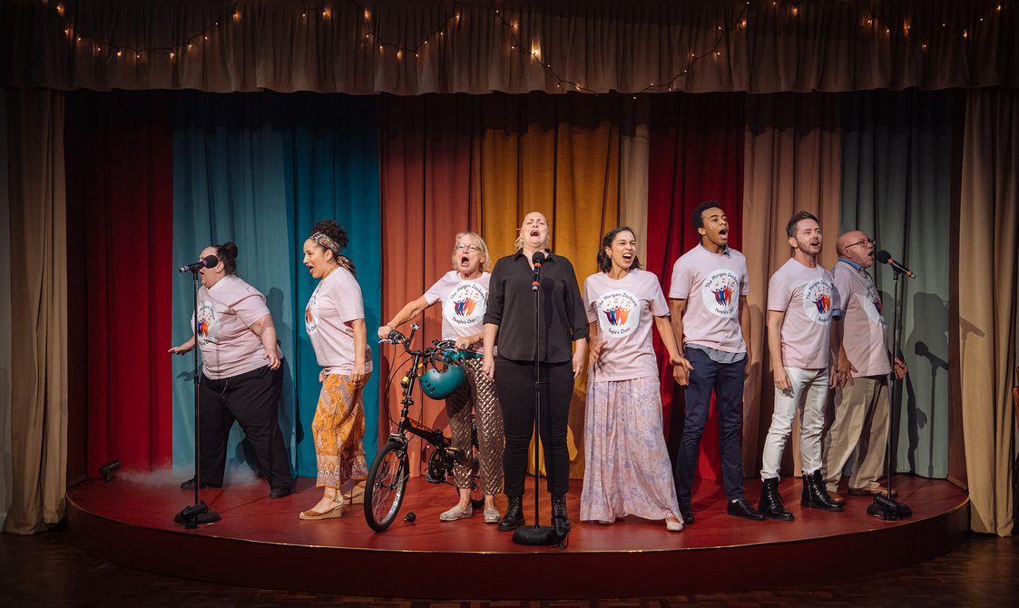 A group of performers are stood in a line on a semi-circular stage, singing passionately. They are all wearing pink t-shirts which say 'The Morgan Jackson People's Choir' and feature colourful figure drawings with musical notes, expect a blonde woman in the centre, who is in all black. The woman to her right is stood next to a bicycle, holding the handlebars which have a teal helmet hanging from them. The backdrop is colourful curtains with fairy lights strewn across the pelmet.