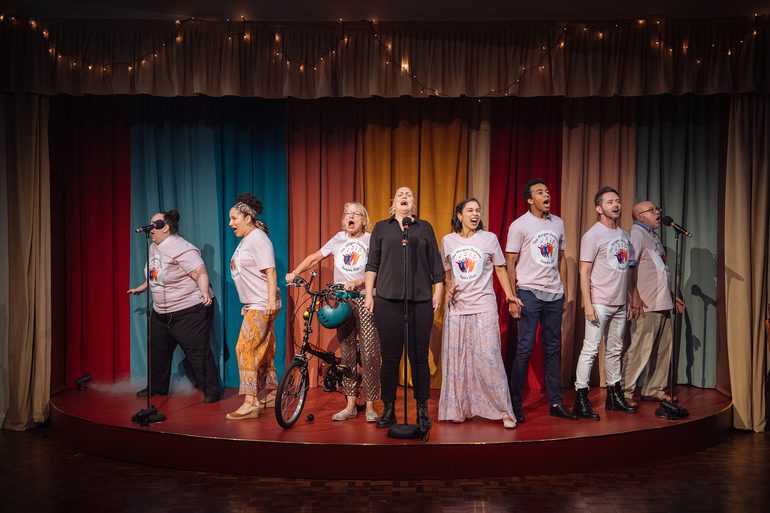 A group of performers are stood in a line on a semi-circular stage, singing passionately. They are all wearing pink t-shirts which say 'The Morgan Jackson People's Choir' and feature colourful figure drawings with musical notes, expect a blonde woman in the centre, who is in all black. The woman to her right is stood next to a bicycle, holding the handlebars which have a teal helmet hanging from them. The backdrop is colourful curtains with fairy lights strewn across the pelmet.