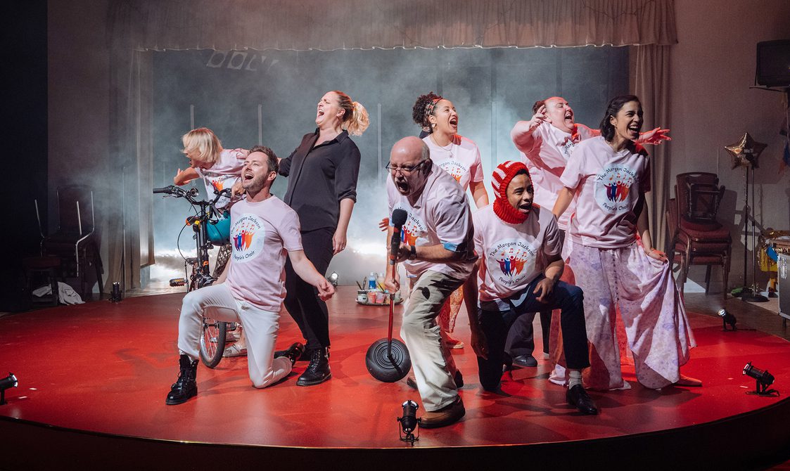 A group of performers are stood on a red, circular stage. They are singing with great intensity. They are all wearing pink t-shirts which say 'The Morgan Jackson People's Choir' and feature colourful figure drawings with musical notes. The woman on the far left is stood next to a bicycle, holding the handlebars which have a teal helmet hanging from them. A faint haze surrounds them.