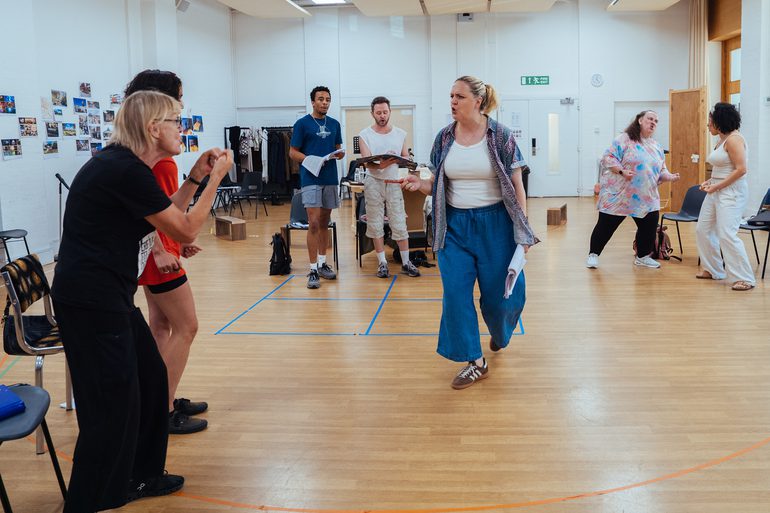 3 pairs of performers are stood around a woman in the centre. She has a somewhat angry expression and is talking to the pair closest to the camera. The next pair are both stood reading their scripts and are singing. The final pair on the right are looking at each other furiously with their clenched fists out in front of them.