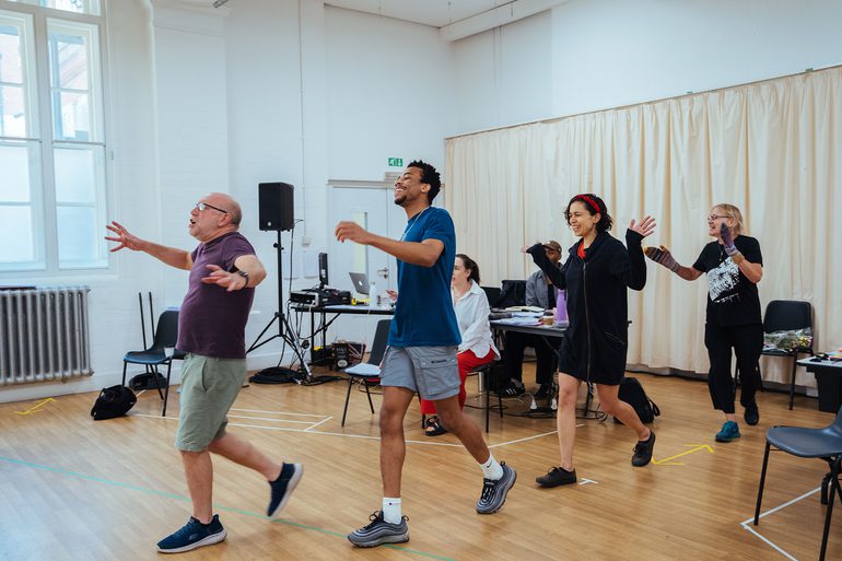 Four performers are marching one in front of the other with their arms swinging. They all have joyful expressions and are singing gloriously.