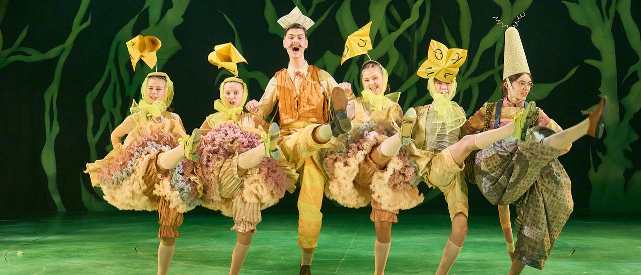 A row of performers kick their legs in unison while linking arms. They are all dressed as yellow ducks. Behind them is a green leafy backdrop. The stage is also a vibrant shade of green.
