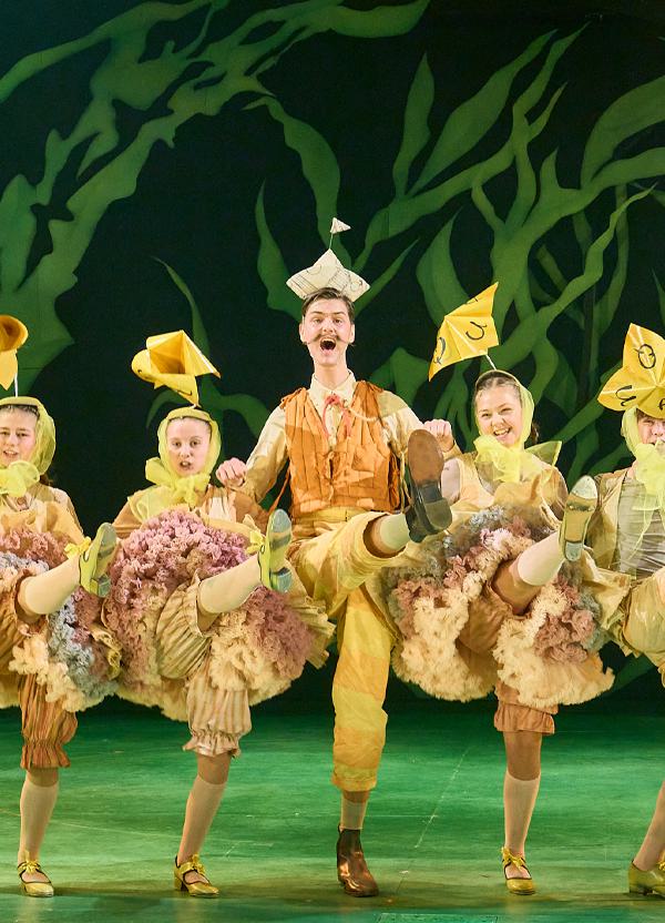 A row of performers kick their legs in unison while linking arms. They are all dressed as yellow ducks. Behind them is a green leafy backdrop. The stage is also a vibrant shade of green.