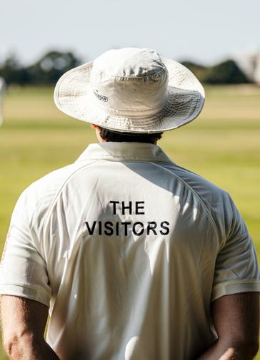 The back of a cricketer in whites with a hat on overlooking a green with cricketers in the distance. On the back of his shirt reads The Visitors in black type.