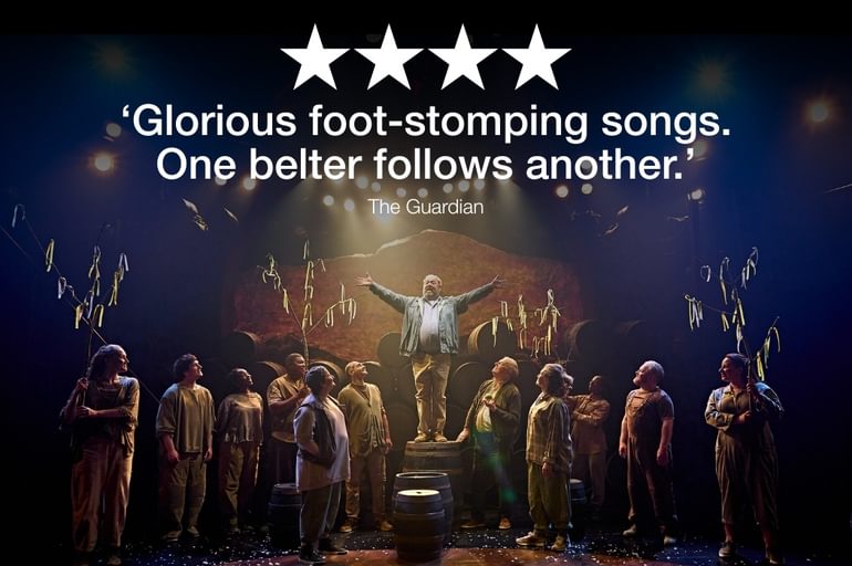 Many characters are gazing wondrously at a man is standing atop a barrel in the centre of the stage. He has his arms wide open. Some of the ensemble are holding tree branches with ribbons tied to them. There are many barrels stacked in the background and others placed amongst the group of people.