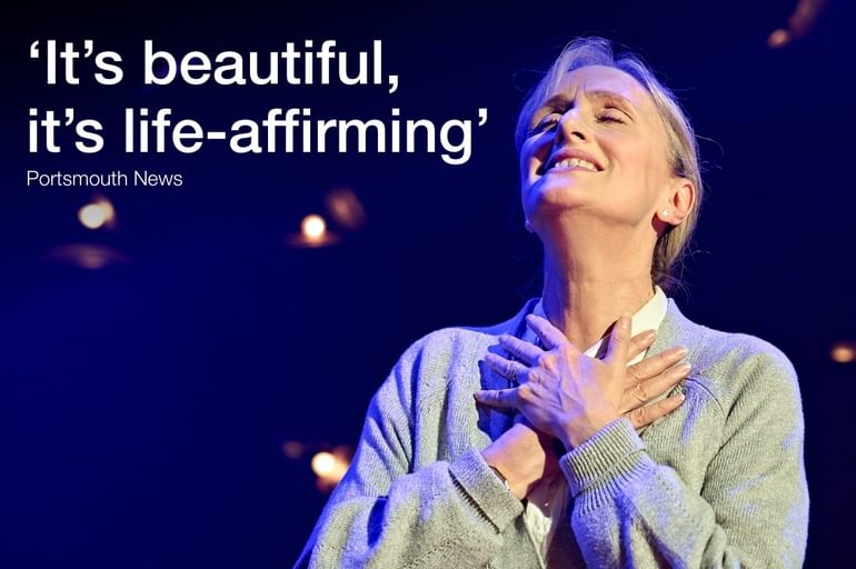 A woman wearing a grey cardigan has her hands protectively placed over one another in the middle of her chest. She has a bright smile on her face as she looks off into the distance.