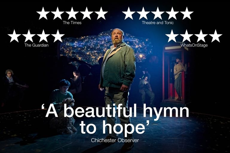 A man is stood upright in the centre of the stage looking off into the distance with a slightly concerned look on his face. Beside him is a shaggy dog puppet being operated by a puppeteer. Further back, there are several people whose faces are illuminated by the light from their mobile phones. There is also young man who is standing casually in a classic-red telephone box. The backdrop is filled with twinkling lights on a deep blue background, reminiscent of a night sky filled with shining stars.
