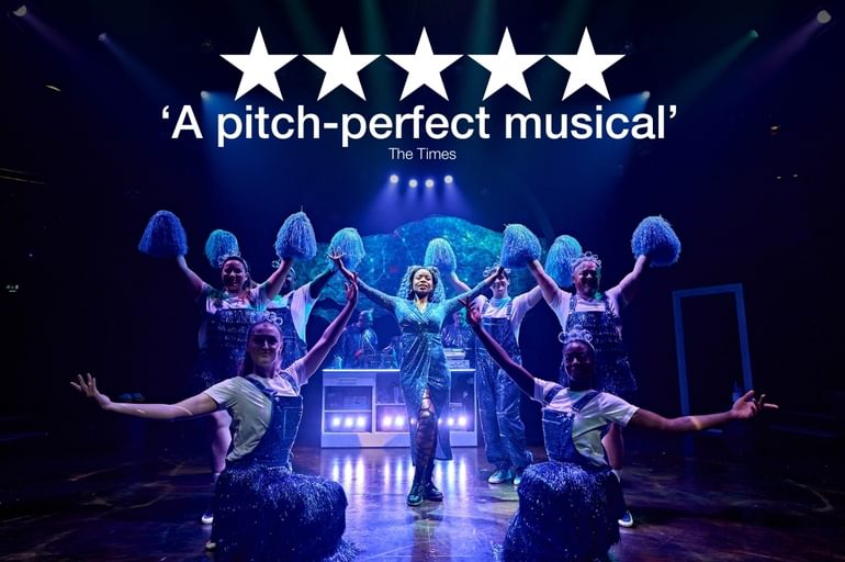 Six dancers are all wearing white t-shirts under sparkling indigo dungarees or pinafores. The two in the front are squatting and have their arms lifted above their heads, while the other four are stood up straight behind them. The standing dancers are holding aloft cheerleader pom-poms. In the centre of this group, a young woman is posing confidently with her arms extended upwards. She is wearing a shimmering wrap dress in a matching colour to the dancers’ costumes with grungy fishnet tights and black combat boots.