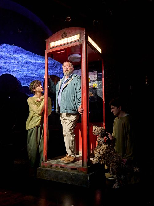 An older man wearing casual clothes is stood inside a worn-down red telephone box. His hands are clasping the doorframe, and he is looking slightly worried. To his right, a young man dressed in an almost threadbare green jumper with a crown of leaves in his hair is gazing up at him inquisitively. On the other side, a shaggy dog puppet operated by a puppeteer is also looking at the man in the phone box.