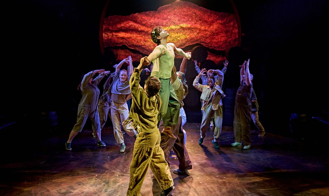 There are several dancers moving fluidly with their arms raised. At the front, a young man is being held up by two people; there is a spotlight shining on him as he looks upward. The backdrop is a fiery sunset on a crumpled, textured background.