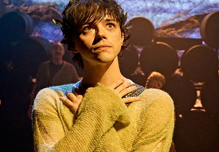 A young man with a neutral expression has his arms crossed over his chest, with his hands resting gently by his neck. He has a crown of leaves amongst his tousled hair and is wearing an almost threadbare green jumper.