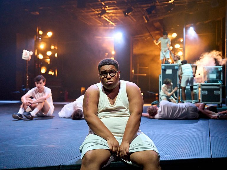 A boy in a white vest and shorts and big, round glasses sits on the edge of the stage. He has a sad expression with his bottom lip protruding. Behind him, several other boys are lying around and others are climbing on flight cases surrounded by haze.