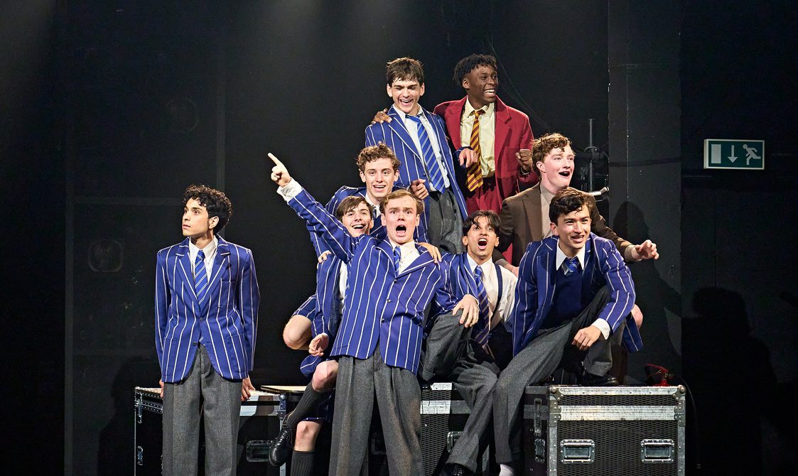 A group of boys dressed in school uniforms sit among several flight cases. They are all laughing and mucking around.