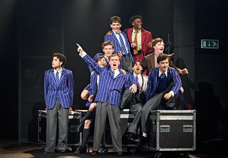 A group of boys dressed in school uniforms sit among several flight cases. They are all laughing and mucking around.