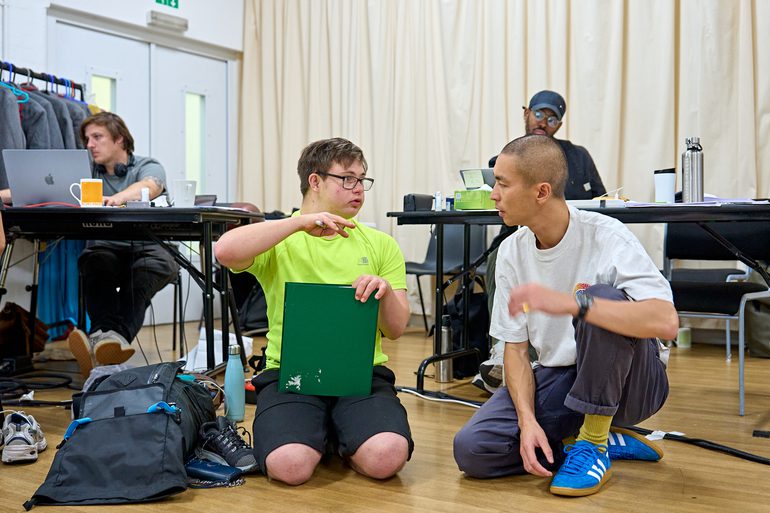 A performer in a bright green t-shirt is knelt on the ground talking to the director who is knelt next to him. The performer is holding a dark green folder and a pen.