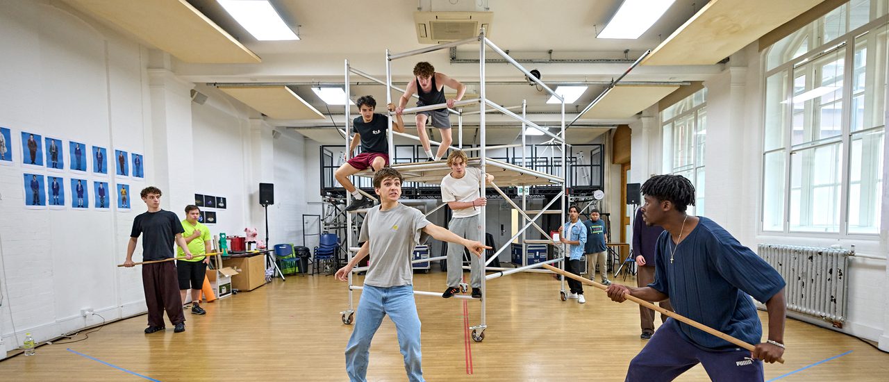 A group of performers are in a rehearsal room. There is a scaffolding rig in the middle of the room which several boys are climbing on. Others are stood around holding sticks. In the front and centre, one boy is pointing to the boy with a stick on the right. He looks panicked.