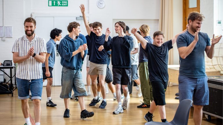 A group of performers in casual clothes are moving around a rehearsal room. Some are clapping happily and others have their hands raised above their heads.