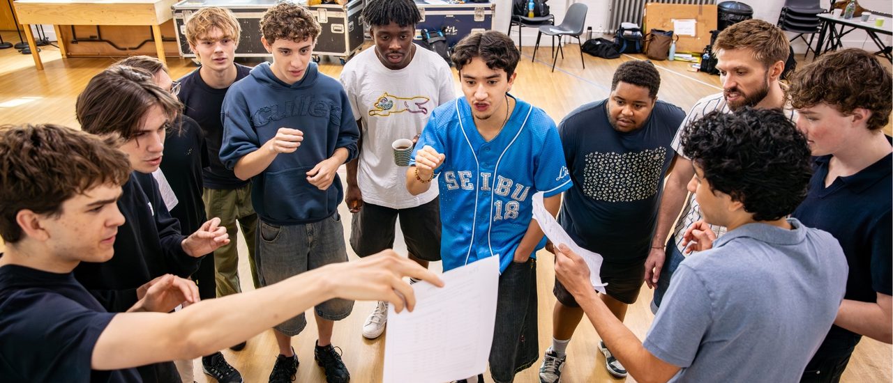 A large group of performers are gathered reading from a piece of paper being held up by the boy on the left. They have focussed expressions.