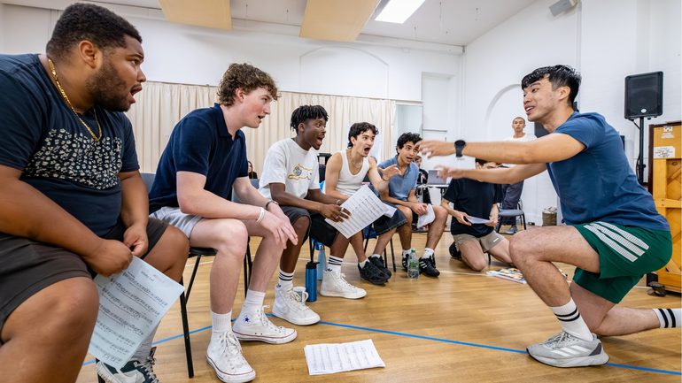 A group of boys are sat in a row singing intensely. They are all facing the musical director (Chris Poon) who is knelt in front of them conducting.