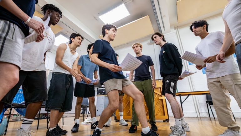 A group of boys are stood close together in a semi circle, with one performer stood in front of them. They are all looking at him. Some are holding sheet music and they all have their lips pursed as if they are about to burst into song.