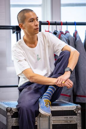 A man (Anthony Lau) is sat casually with on a large black stage case. Behind him is a clothing rail with grey blazers hanging.
