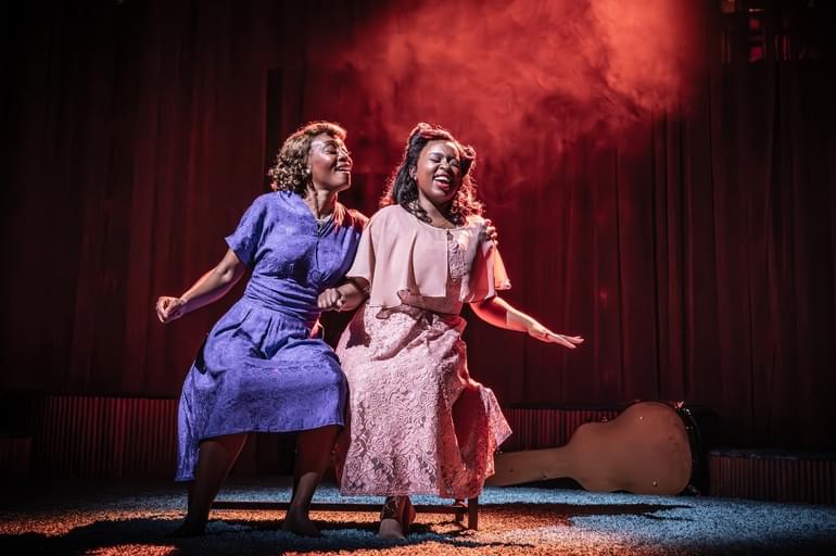 Two Black women are sat down. They both have their arms out as if dancing and have brilliant smiles on their faces. The lighting on the stage is red and there is a faint haze behind them.