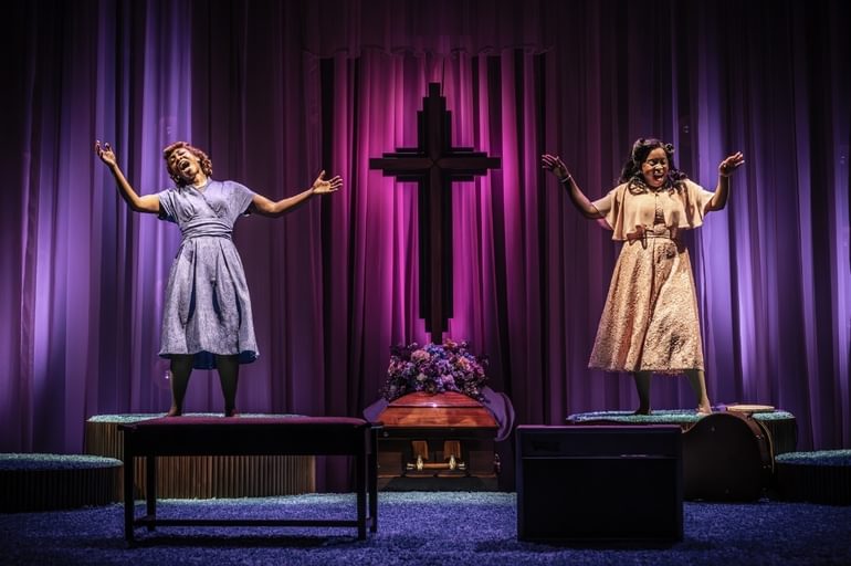 Two Black women are singing passionately with their eyes closed and arms extended freely on two raised platforms either side of a large crucifix with flowers at the base. There are luxurious indigo and magenta curtains flowing behind them.