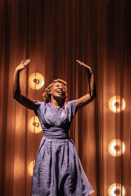 A Black woman is singing with a gleeful expression and her arms raised above her head. The lighting behind her is warm and comforting. Her dress is a muted blue with a subtle circular pattern and her hair is styled with a victory roll.
