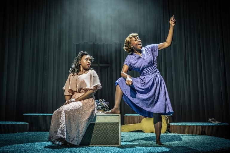 Two Black women are in the centre of a stage covered with teal shag carpet. The younger woman on the left is sat on a box with her hands in her lap, looking over her shoulder at the older woman on the right who is confidently stood with one foot propped up on the box. She is singing powerfully, facing away from the other woman with one arm raised.