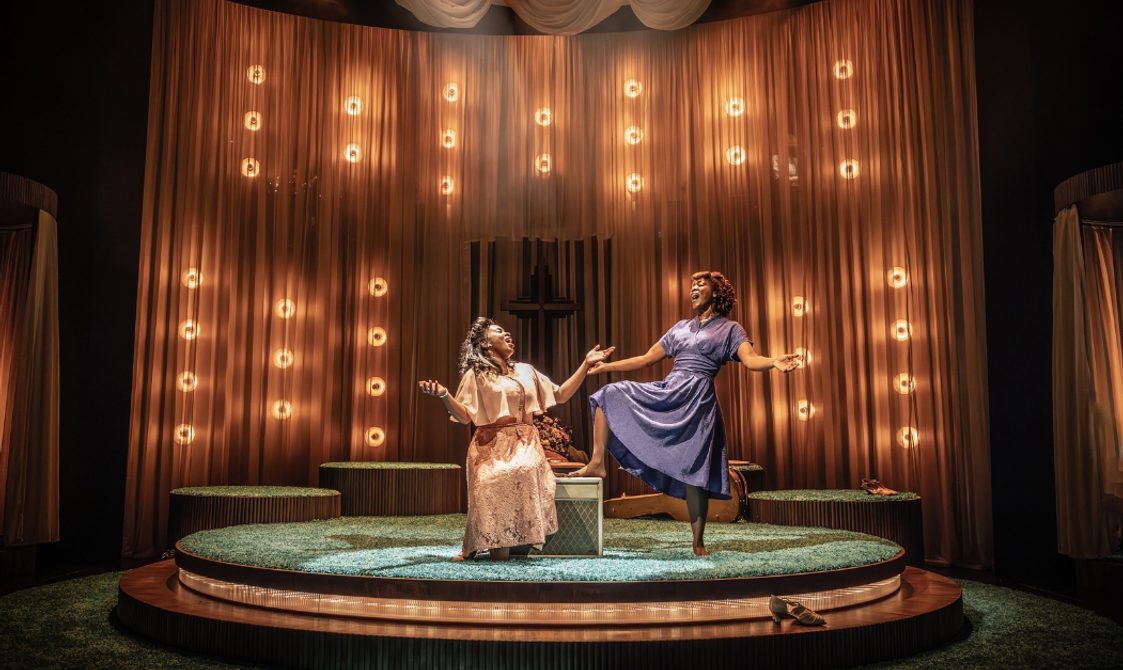 Two Black women are singing joyfully with their arms extended expressively facing each other. The younger woman on the left is sat on a box wearing a pale pink floral dress with a flowing shawl and the older woman on the right who is confidently stood with one foot propped up on the box. She is barefoot and wearing a muted blue knee length dress. Their styling is evocative of the 1940s.