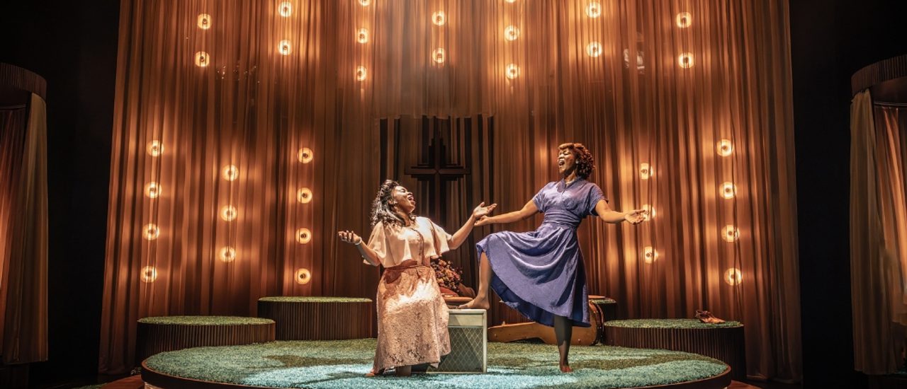 Two Black women are singing joyfully with their arms extended expressively facing each other. The younger woman on the left is sat on a box wearing a pale pink floral dress with a flowing shawl and the older woman on the right who is confidently stood with one foot propped up on the box. She is barefoot and wearing a muted blue knee length dress. Their styling is evocative of the 1940s.