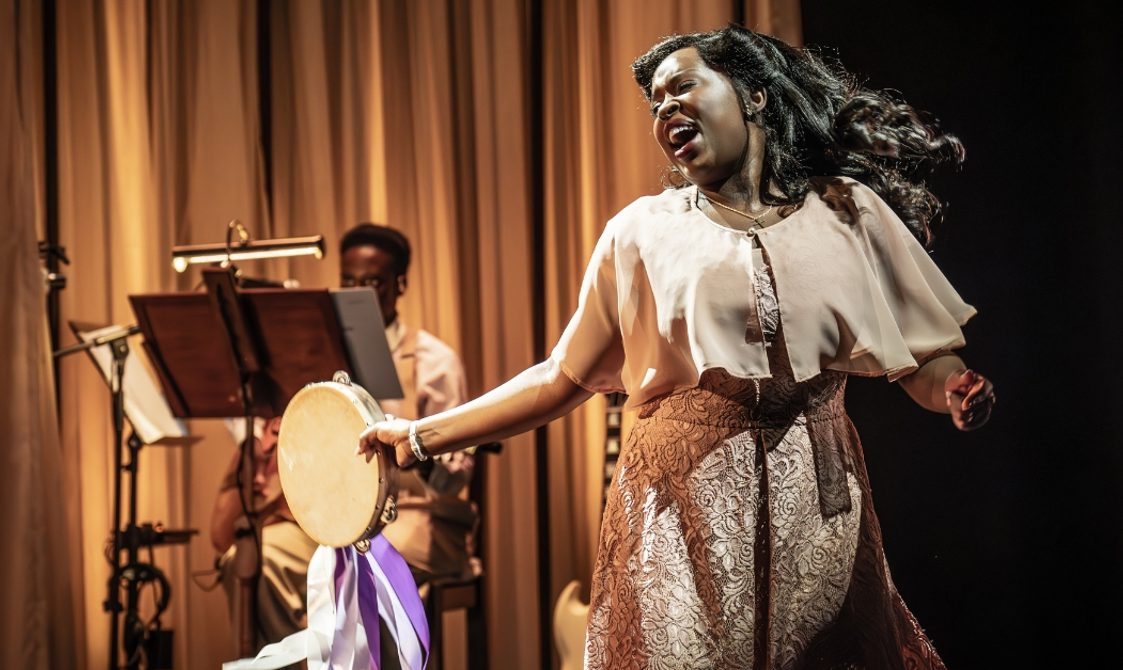 A young Black woman is singing passionately with a tambourine that has cascading purple and white ribbons in her hand. Her hair is in 1940s style victory rolls, with a flowing pale pink shawl over a floral jacquard dress in the same colour and a simple crucifix around her neck. There is a musician playing the guitar in the background with a music stand in front of them.
