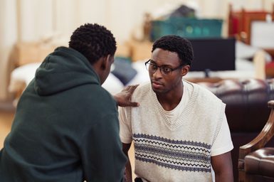 Two performers are crouched down together. The young man on the left has his back to the camera and holds his hand out to the shoulder of the other young man, who looks at him with a slight frown, as though worried or sad.