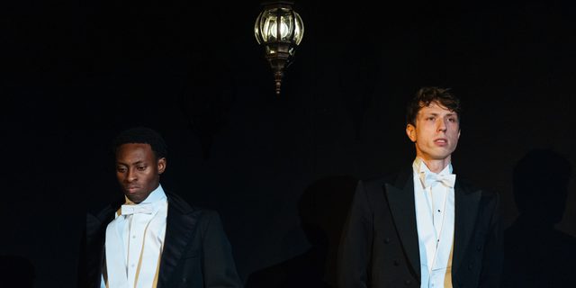 Two young men in dinner jackets stand side by side. The man on the left looks at the ground in annoyance. The man on the right looks out in front of him with an awkward expression.